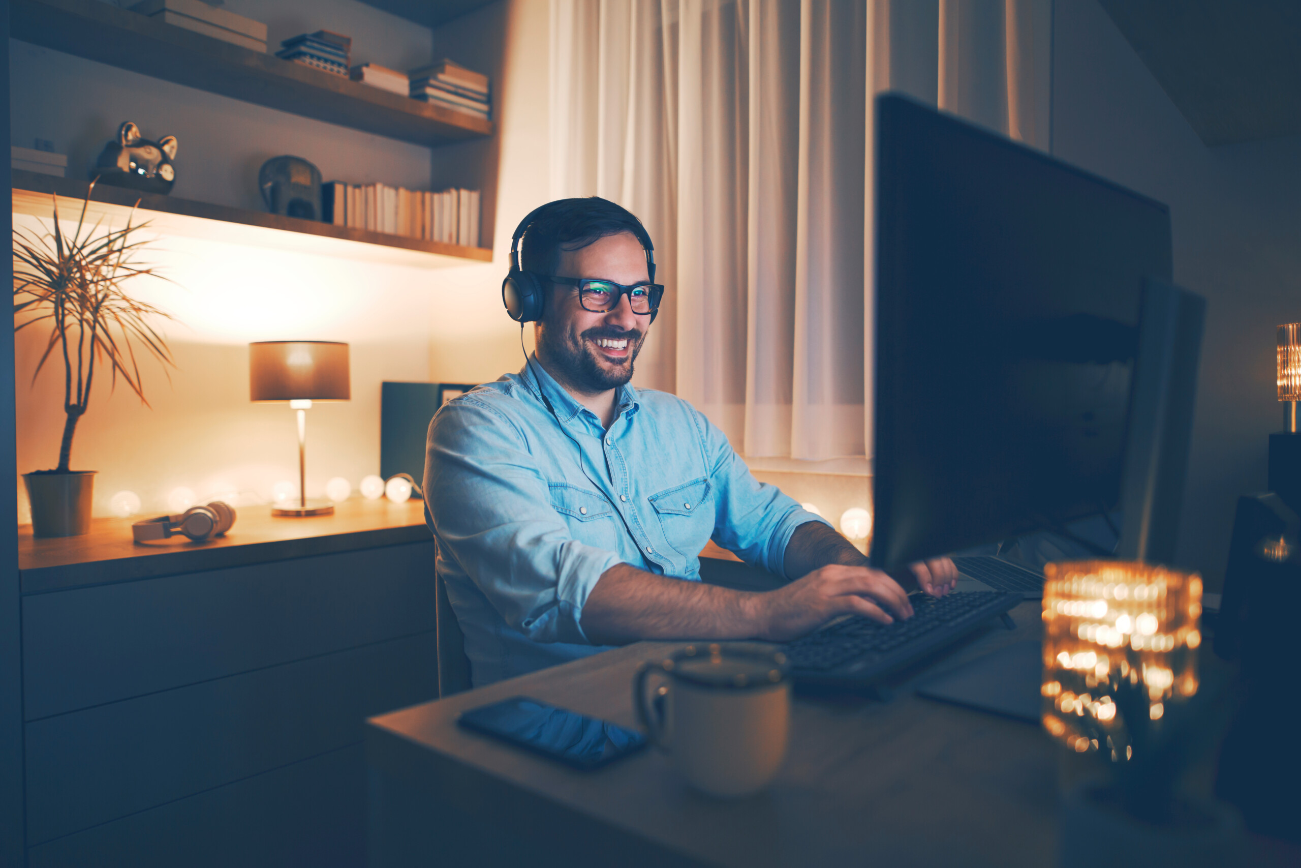 Modern young man working remotely from home at night. He is using headphones.